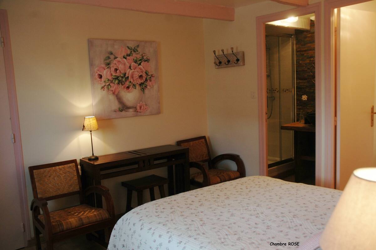 A bedroom with a bed, a desk, two chairs, a lamp, a painting of flowers, and a bathroom door.