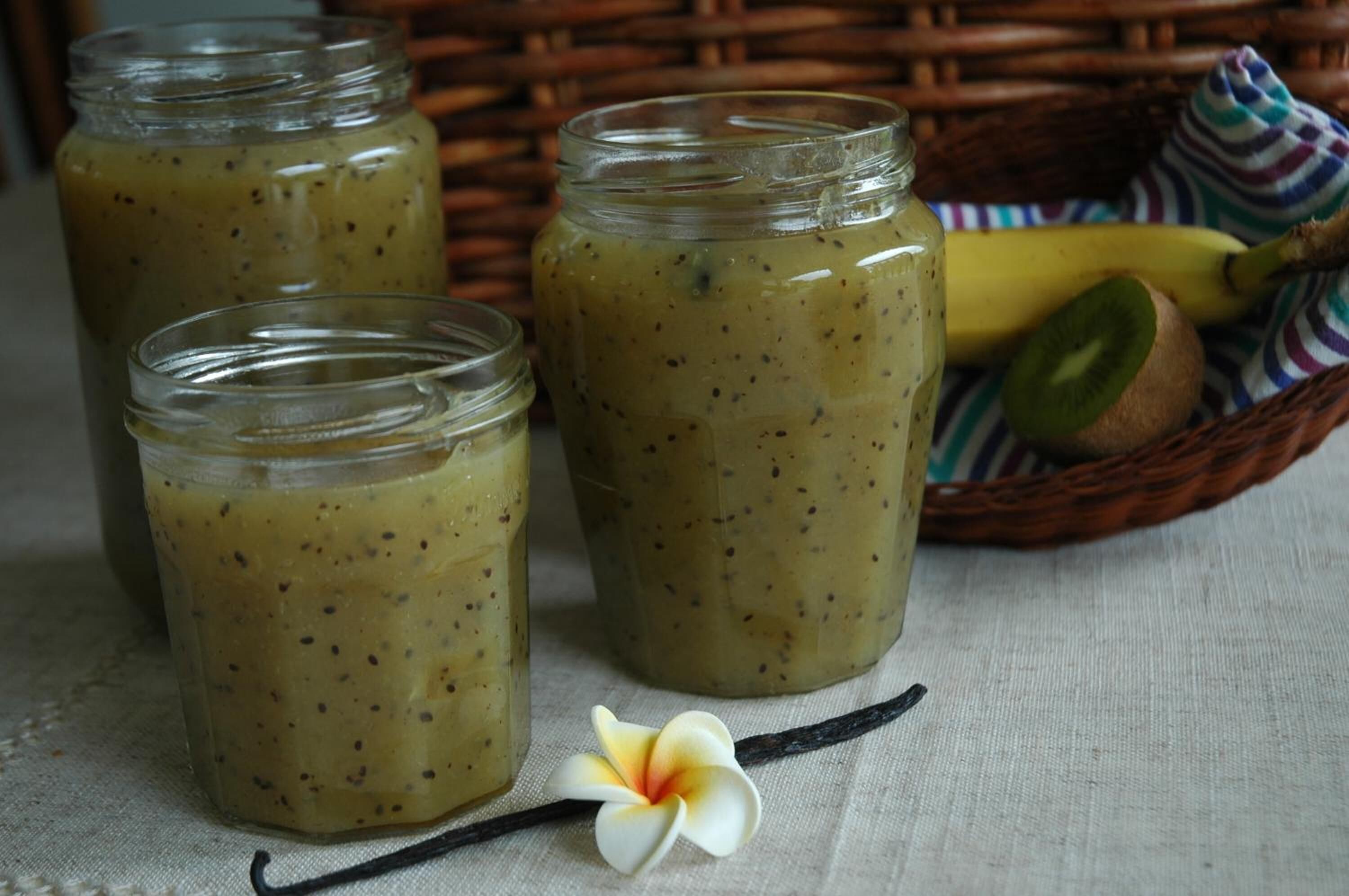 Three glass jars with a greenish mixture, a banana, kiwi, and a vanilla pod on a cloth.