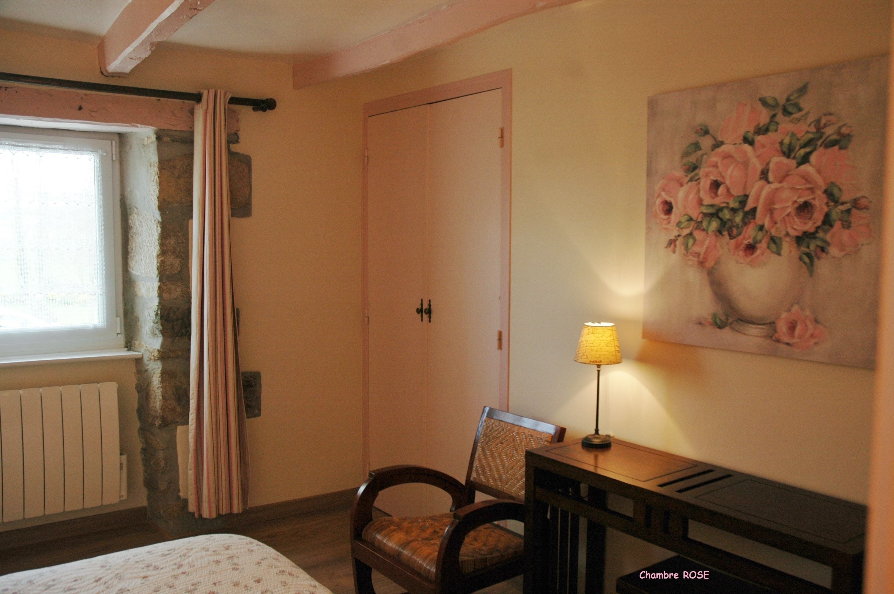 A bedroom with a bed, a chair, a desk with a lamp, a painting of flowers, and a door.