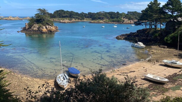 Escena costera con barcos amarrados en una playa de arena, rodeada de afloramientos rocosos y árboles.