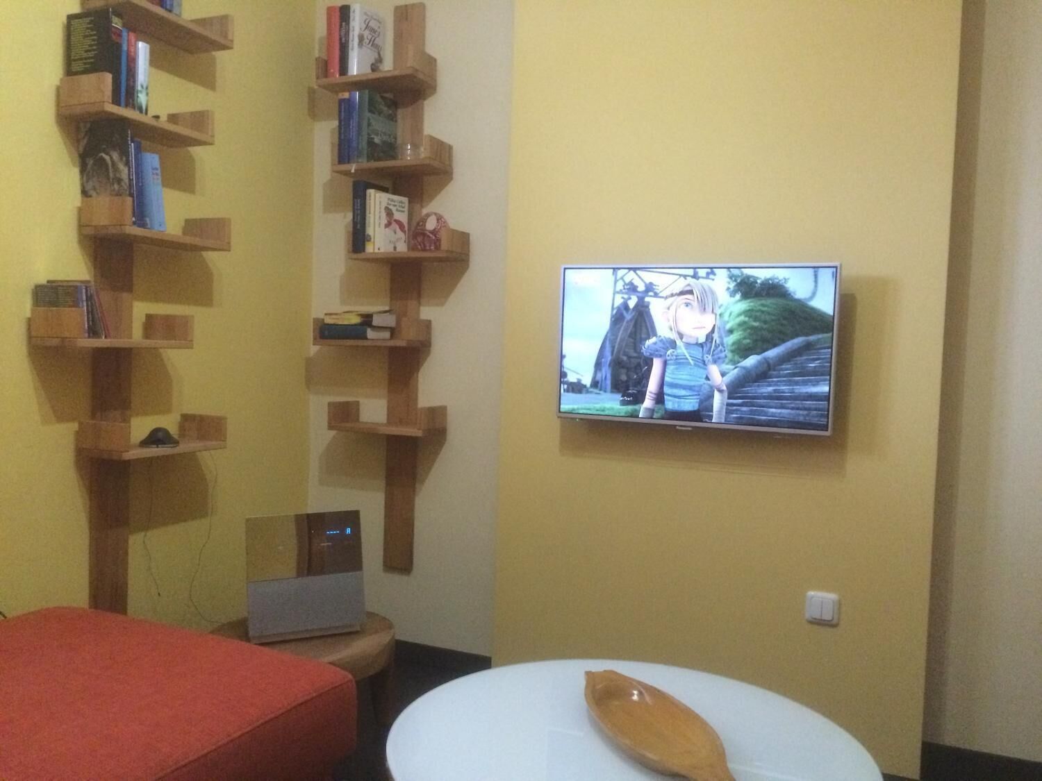 A room with a TV mounted on the wall displaying a scene from a movie, wooden bookshelves filled with books, a small desk with a computer, and a red couch.