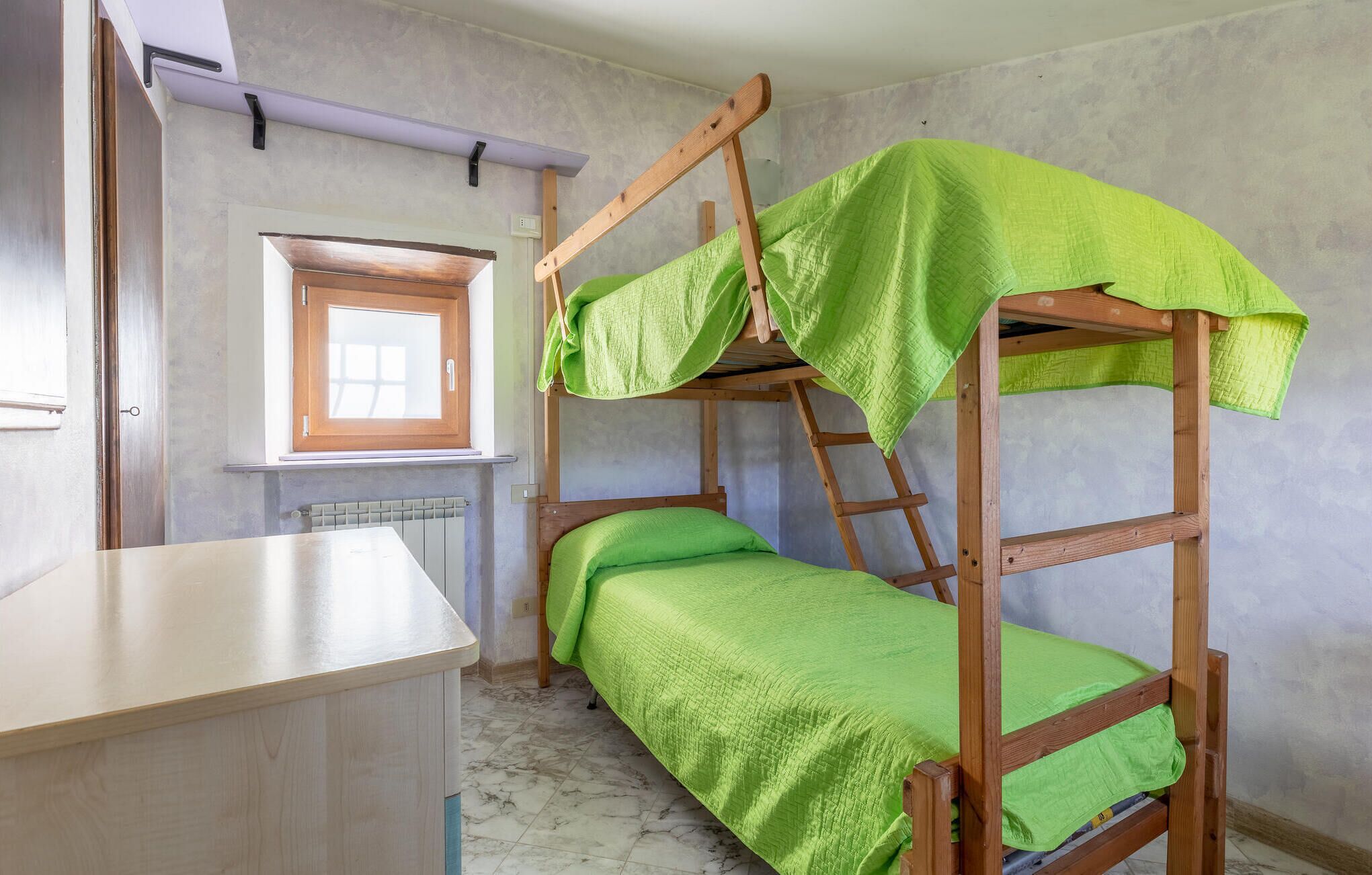 Un lit superposé avec une literie verte, un bureau en bois, une fenêtre avec un cadre en bois et un radiateur posé sur le sol.