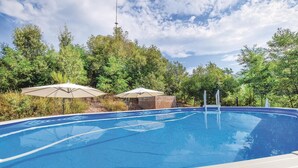A swimming pool with a clear blue water surface, surrounded by lush greenery and trees. There are two umbrellas providing shade.