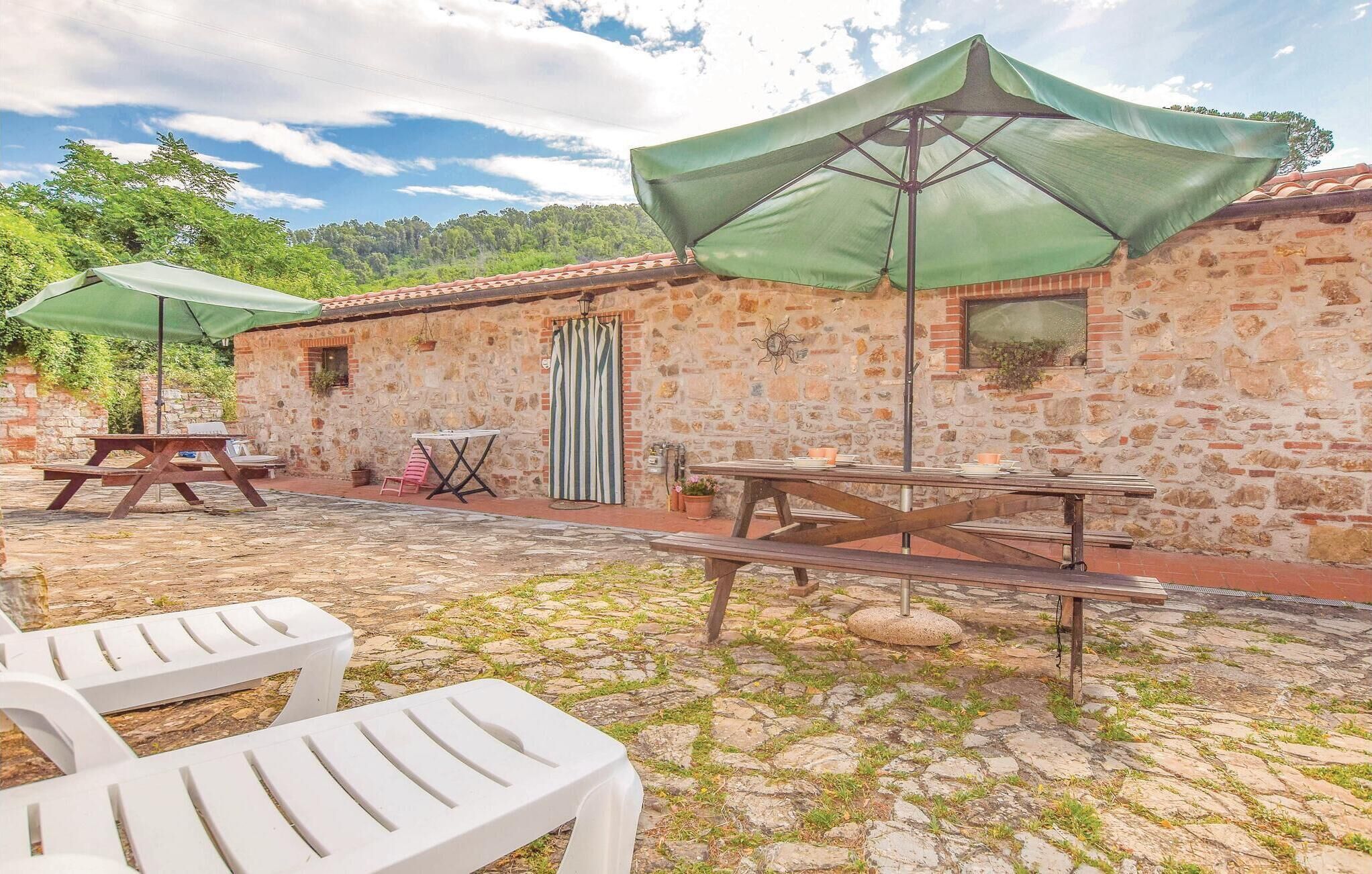 An outdoor patio area with stone flooring, picnic tables, and chairs, surrounded by stone walls and green umbrellas.