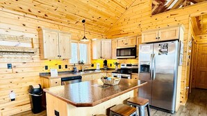 Interior de una cabaña de madera con cocina que incluye una isla central, taburetes de bar, un refrigerador de acero inoxidable y armarios de madera.