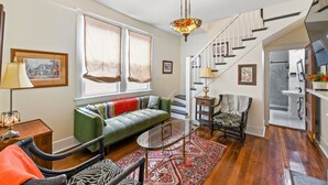 Ein Wohnzimmer mit einem grünen Sofa, einem Glastisch und einem gemusterten Teppich. Eine Treppe mit weißer Geländer führt zu einem Badezimmer.