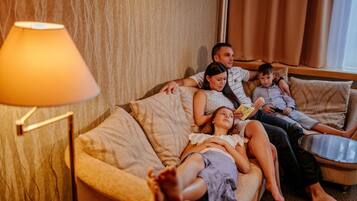 Une famille de quatre personnes est assise sur un canapé beige dans un salon, avec une lampe et une vue sur des rideaux.
