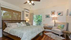 Un dormitorio con una cama de madera, ventilador de techo, ventana con persianas, mesita de noche con lámpara, una silla y una alfombra.