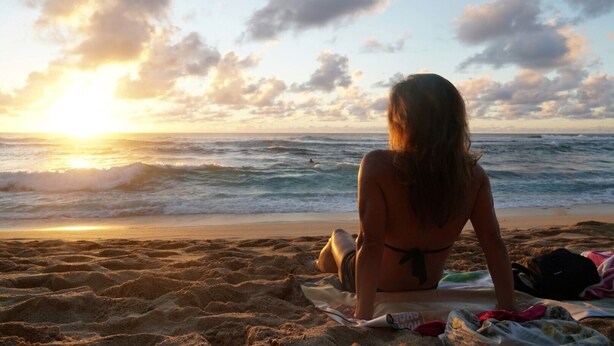 Una persona sentada sobre una toalla de playa mirando el océano al atardecer.