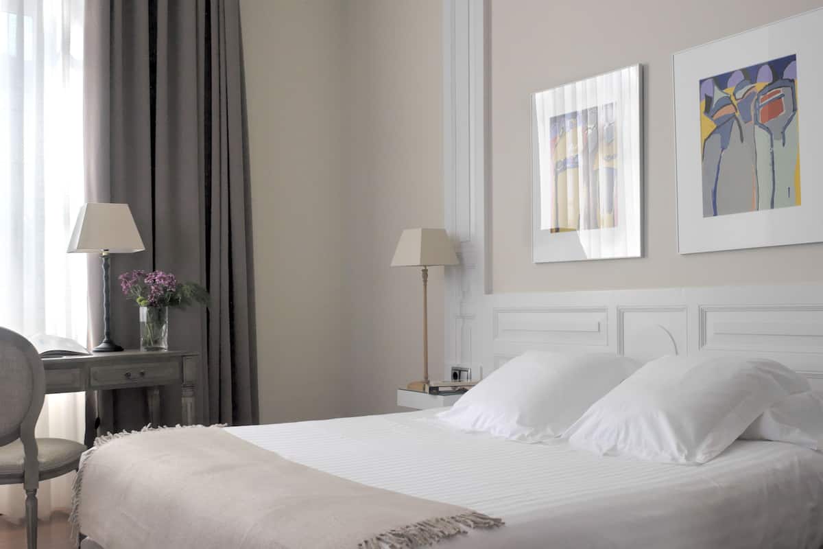 Een opgemaakt bed met witte lakens, een nachtkastje met een lamp en een stoel.