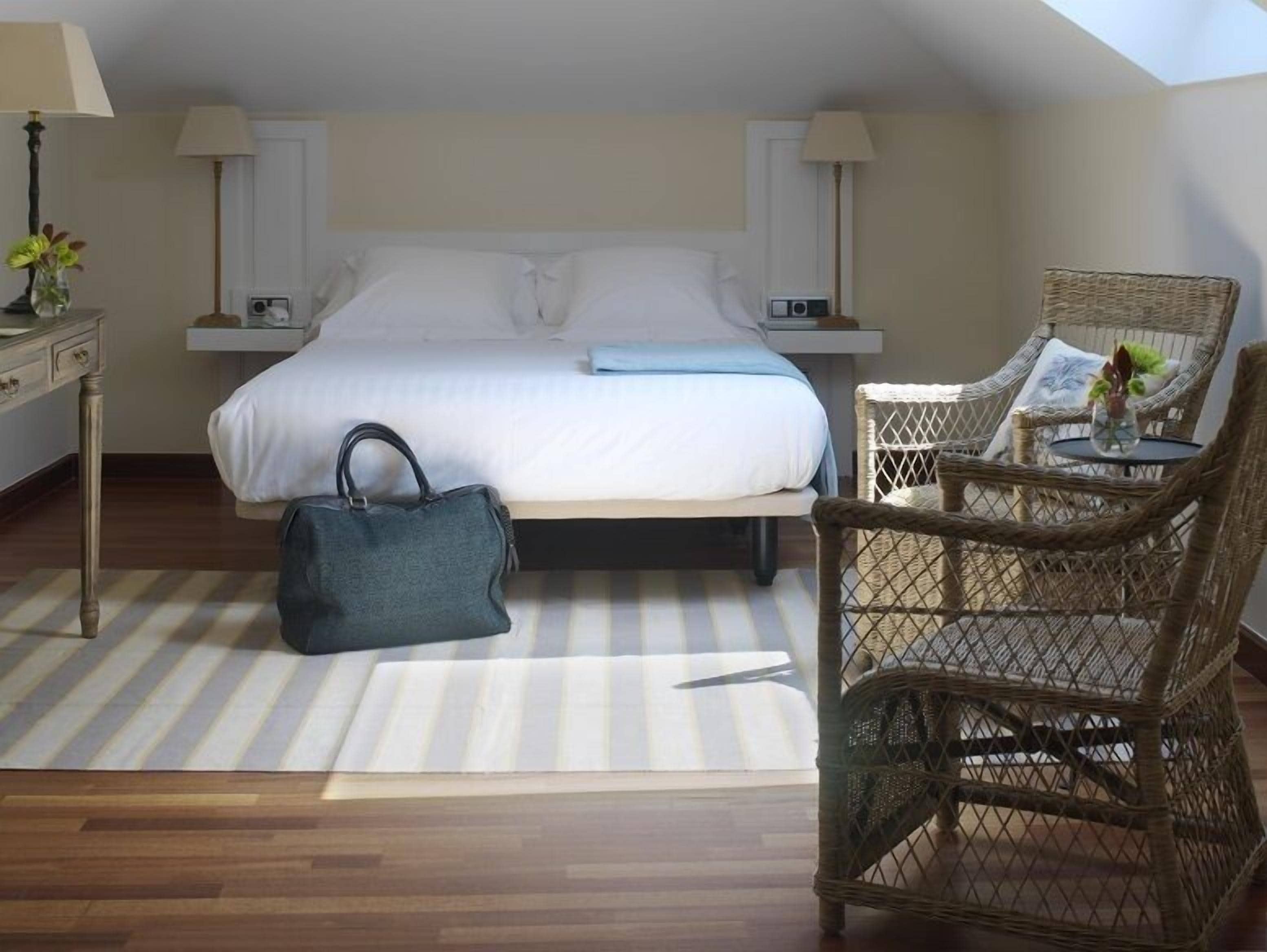 Habitación de hotel con cama, una silla de mimbre, mesita de noche, lámpara y bolso de mano.