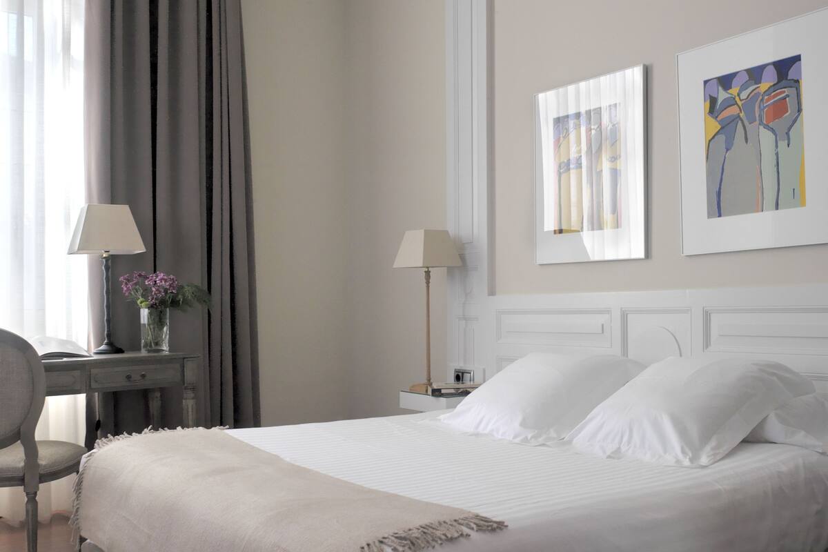 Een opgemaakt bed met witte lakens, een nachtkastje met een lamp en een stoel.