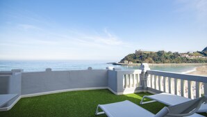 Un balcon agrémenté de gazon synthétique, de chaises longues blanches et offrant une vue sur la mer et une colline parsemée de bâtiments.