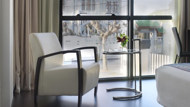 Una habitación de hotel moderna con un ventanal grande, una silla blanca, una mesita con un florero y una cama.