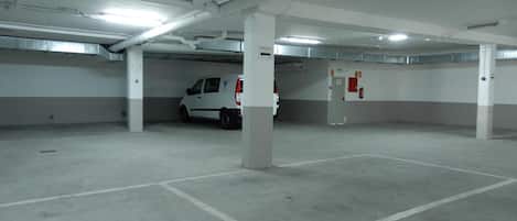 Un estacionamiento con pilares blancos, una camioneta blanca estacionada en uno de los espacios, y un extintor colgado en la pared.