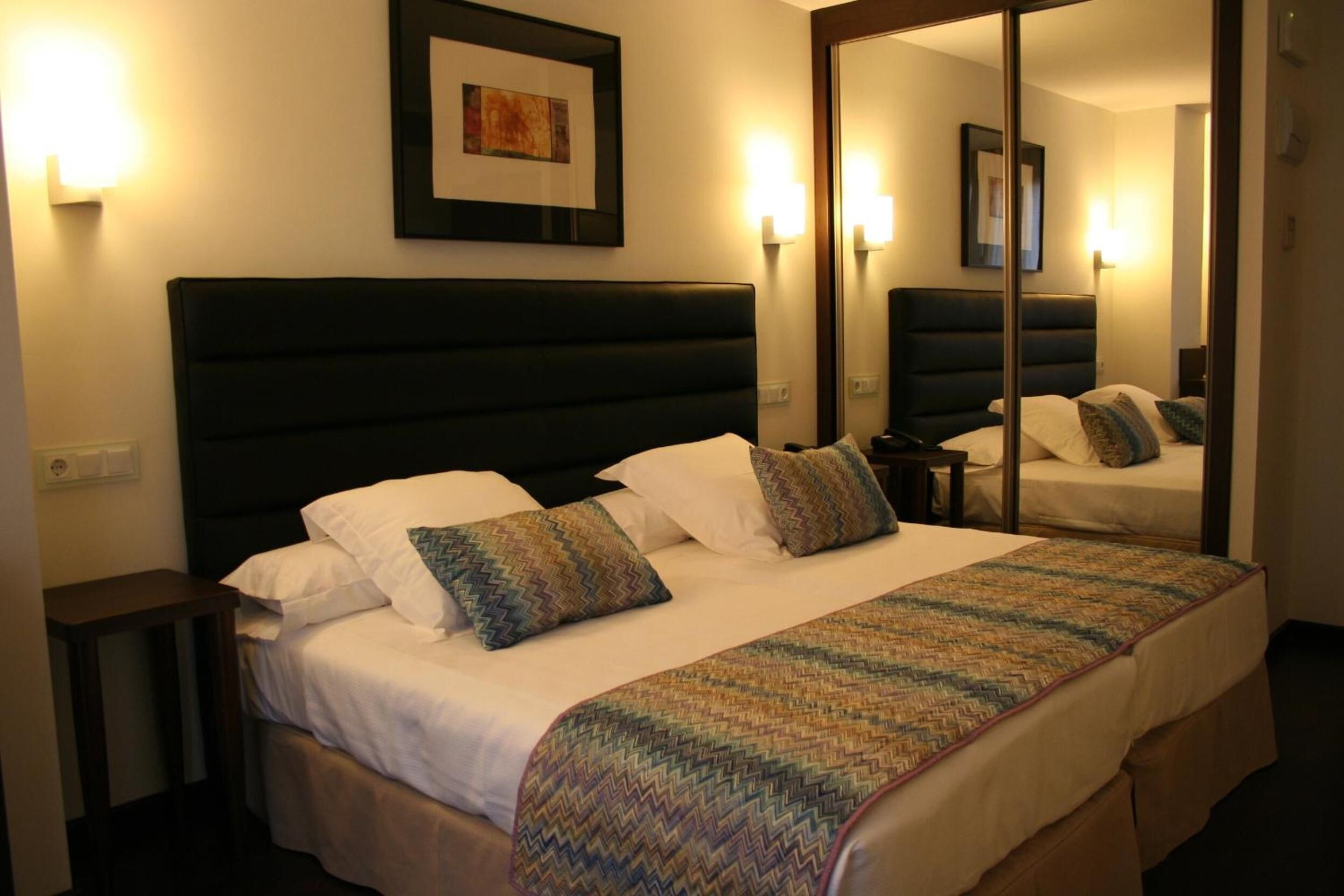 A hotel room with a large bed, a bedside table, a mirror, and wall-mounted lights.