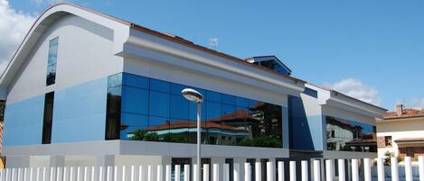 A modern building with a white and blue facade, large windows, and a white fence in the foreground.