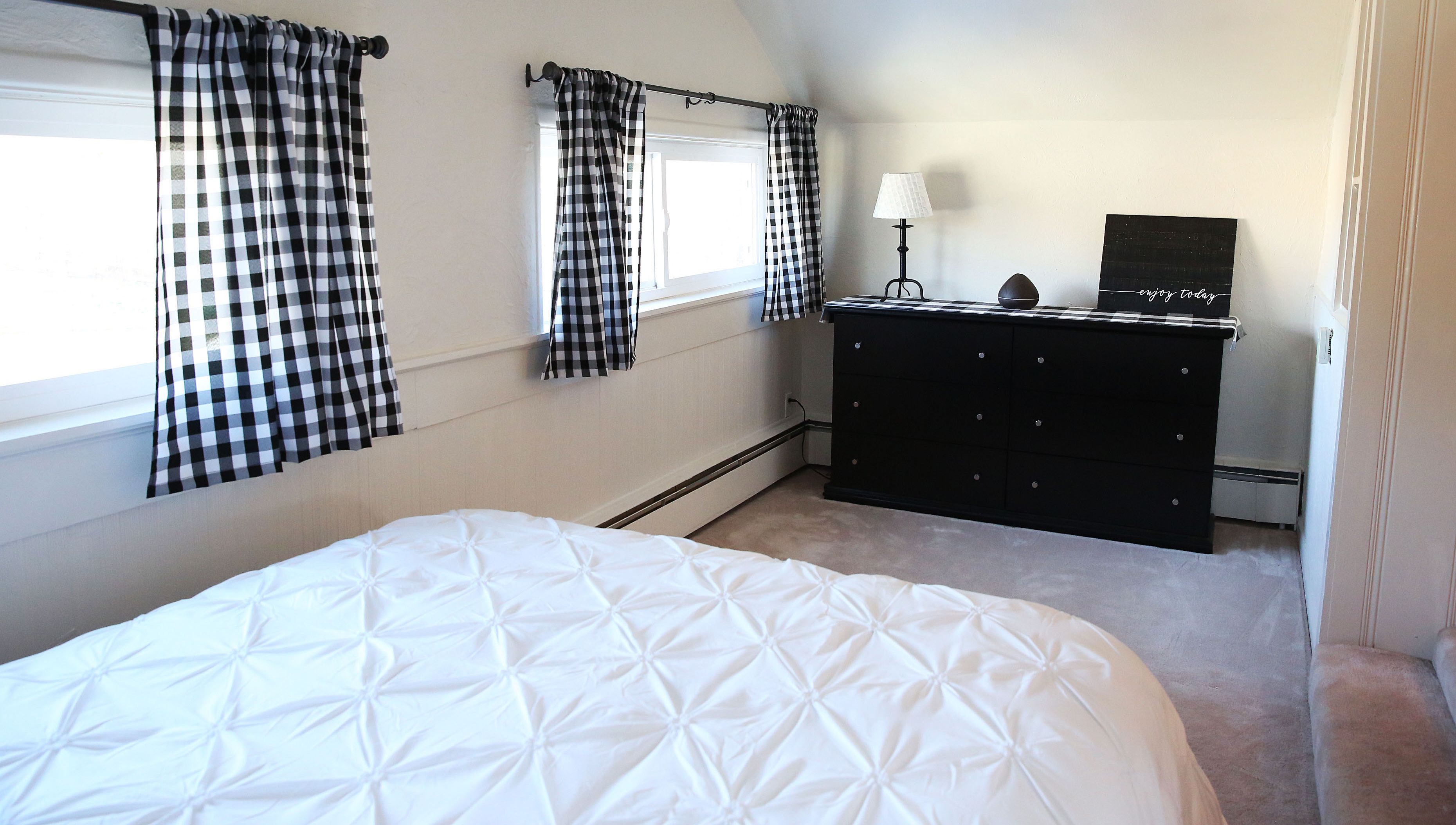Un dormitorio con cama, dos ventanas con cortinas a cuadros, un aparador y una lámpara de pared.