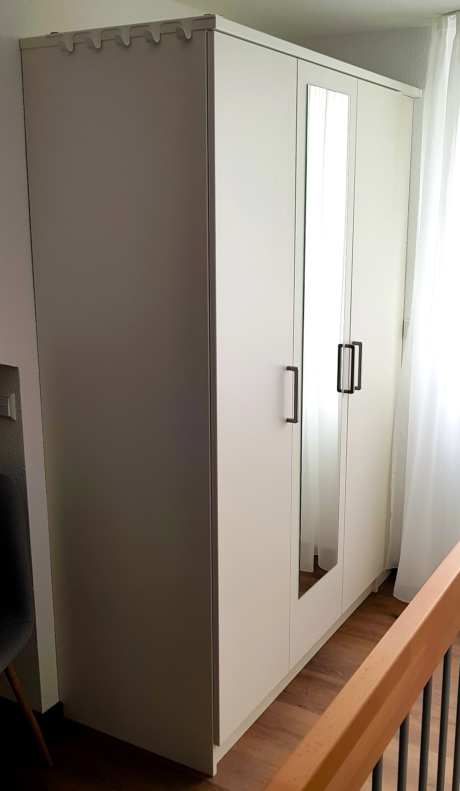 A white wardrobe with a mirrored door and a handle, located in a room with a wooden floor and a white railing.