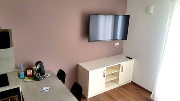 A small, modern room with a flat-screen TV mounted on the wall, a kitchenette area with a microwave, and a dining table with chairs.
