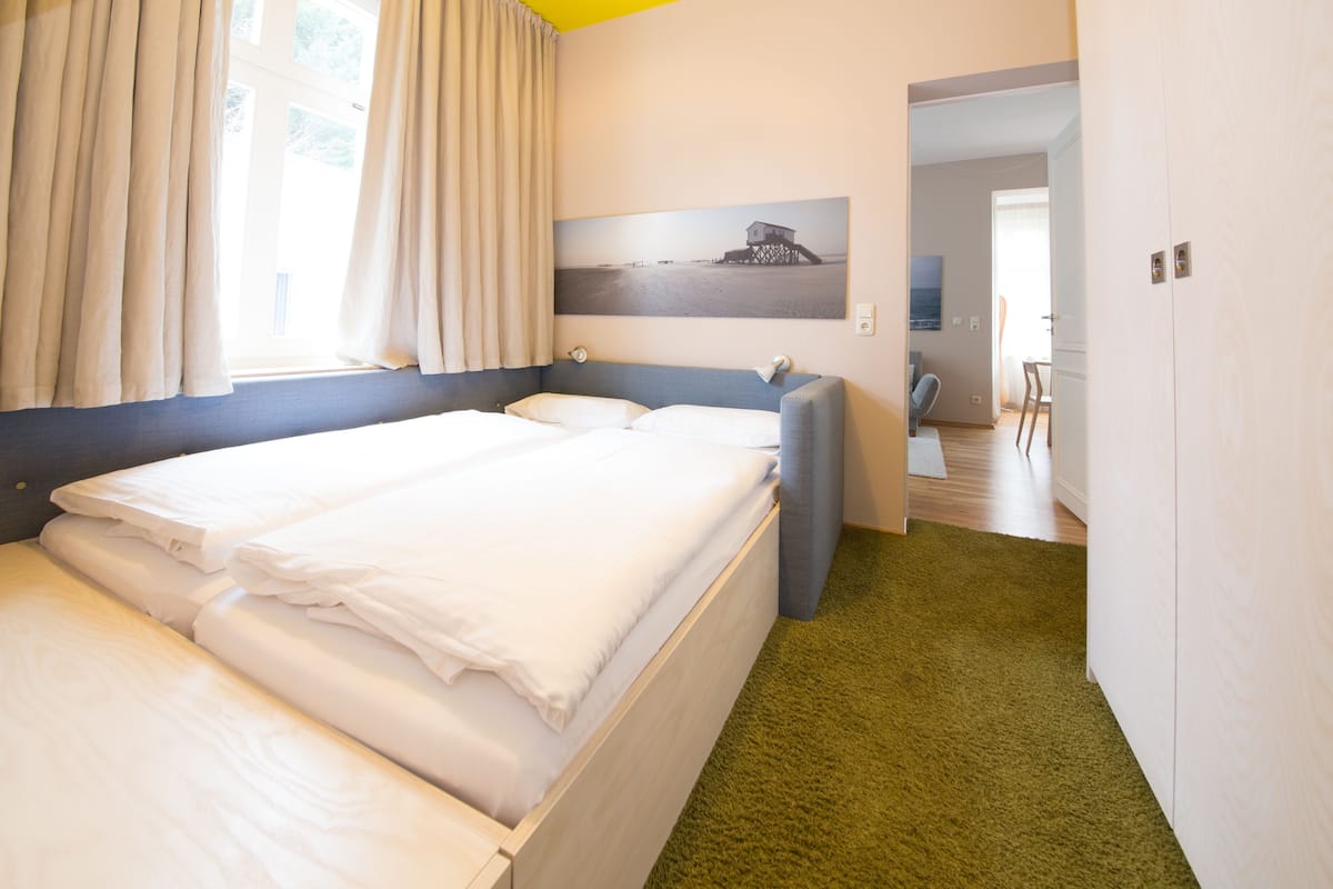 A hotel room with a bed, a desk, and a chair. There is a window with curtains, a painting on the wall, and a green carpet.