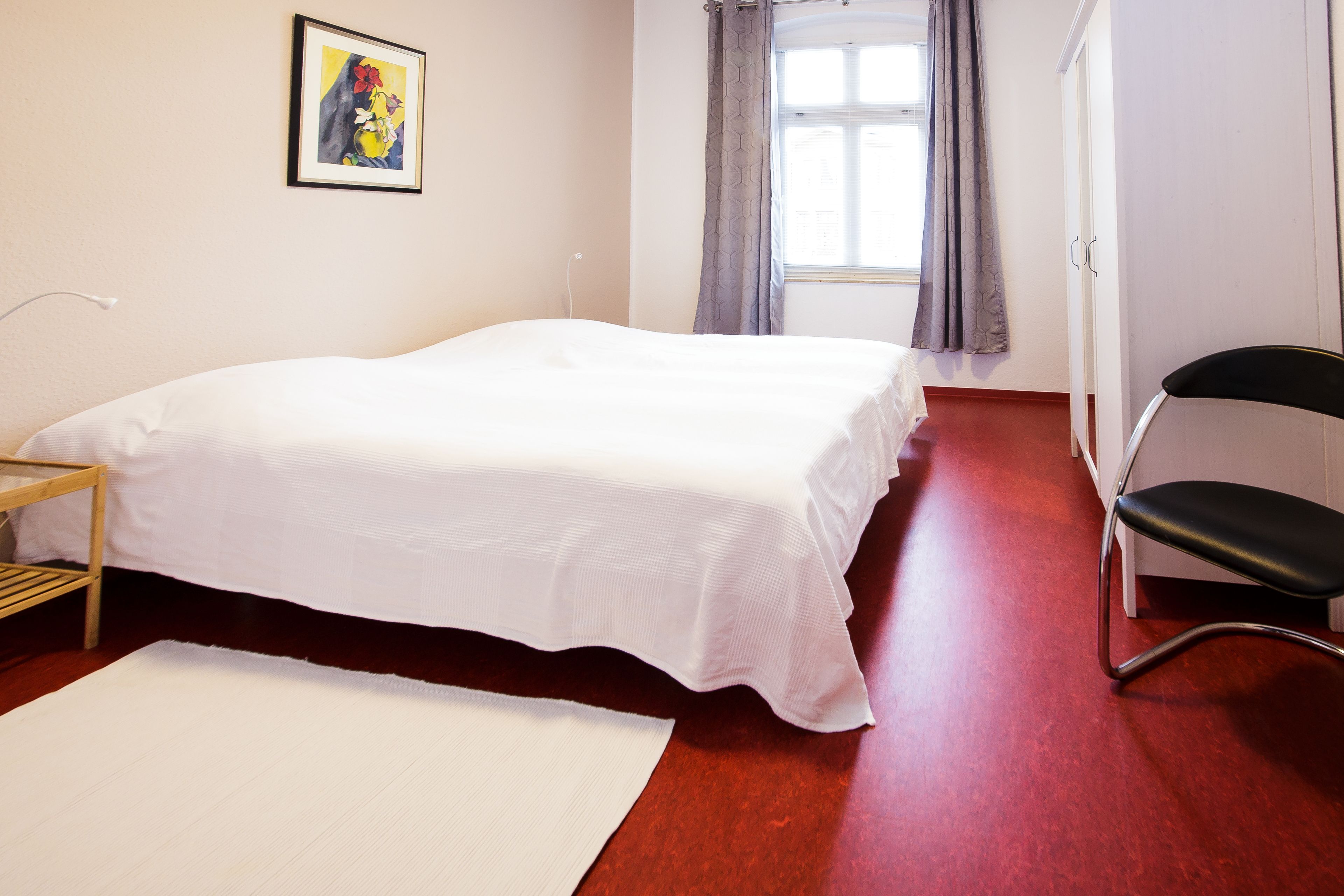 A bedroom with a bed, a chair, a window with curtains, and a painting on the wall.