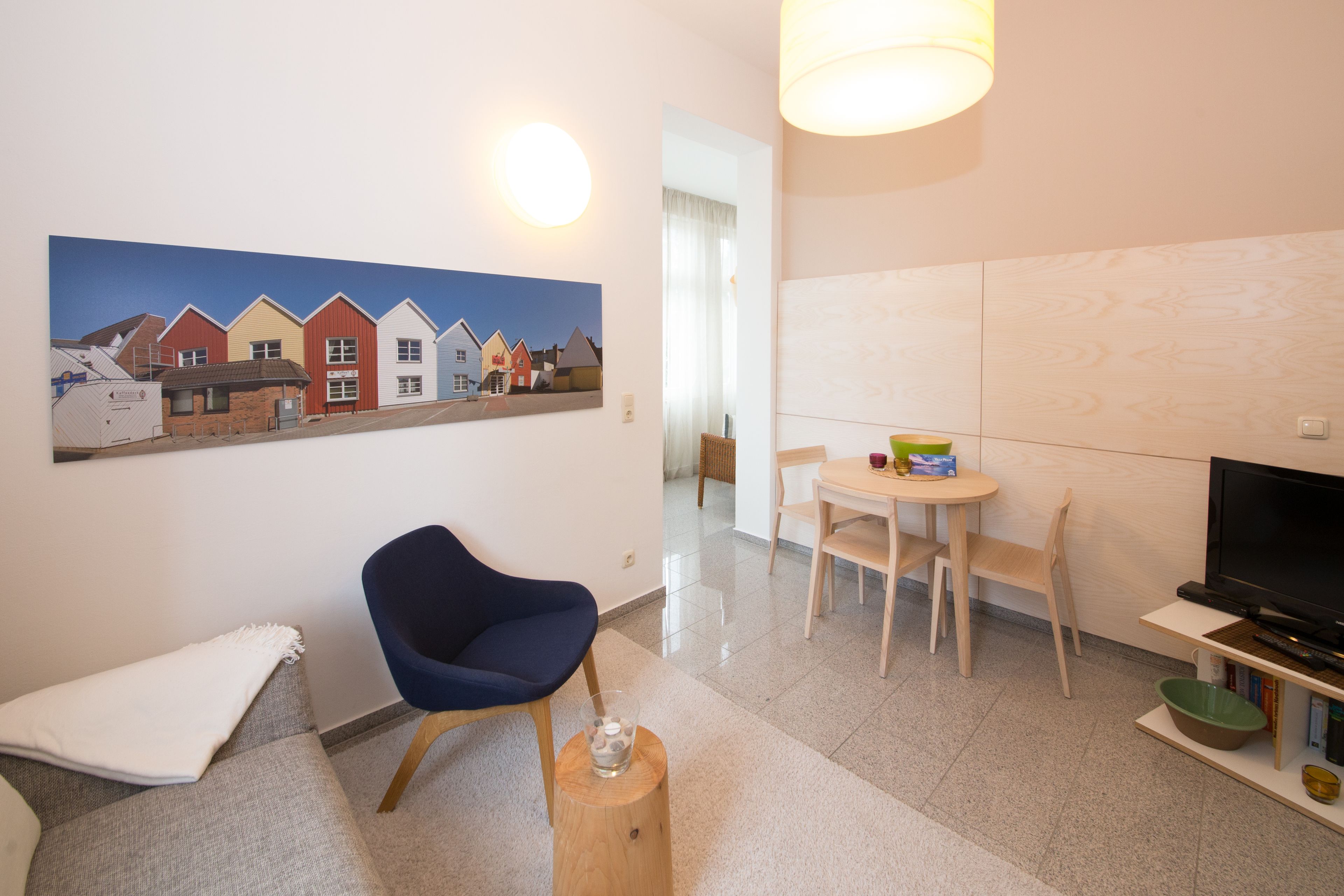 A modern living room with a blue chair, a dining table with chairs, a TV, and a painting of houses on the wall.
