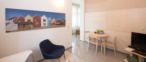 A modern living room with a blue chair, a dining table with chairs, a TV, and a painting of houses on the wall.