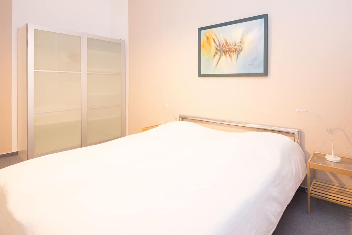 A hotel room with a bed, a bedside table with a lamp, a wardrobe, and a framed abstract painting on the wall.
