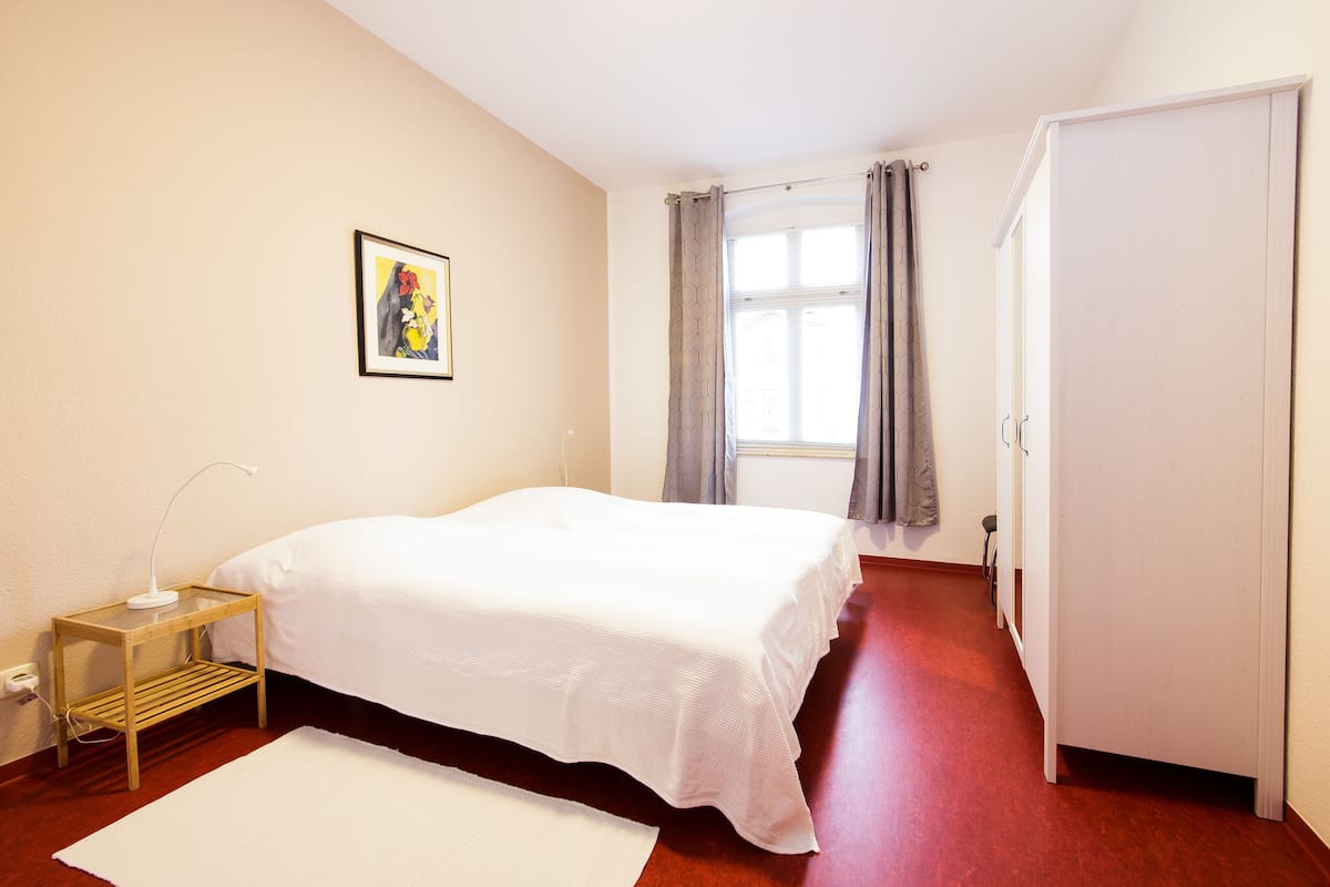 A bedroom with a bed, a bedside table, a lamp, a window with curtains, and a door.