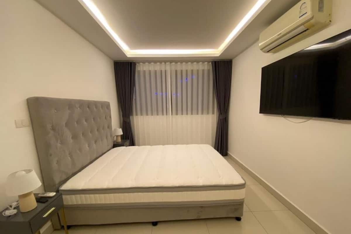 A bedroom with a bed, bedside table, lamp, air conditioning unit, and a TV mounted on the wall.
