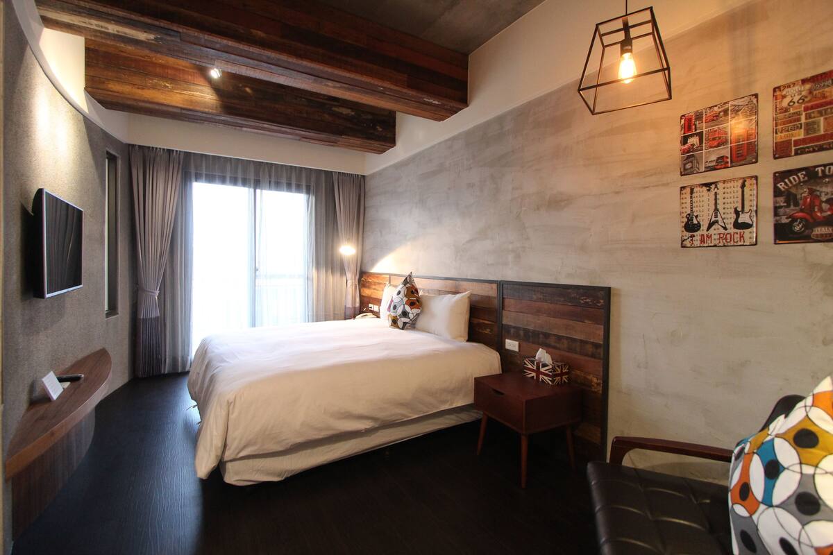 A hotel room with a large bed, a wooden headboard, a bedside table with a lamp, a flat-screen TV mounted on the wall, and a window with curtains.