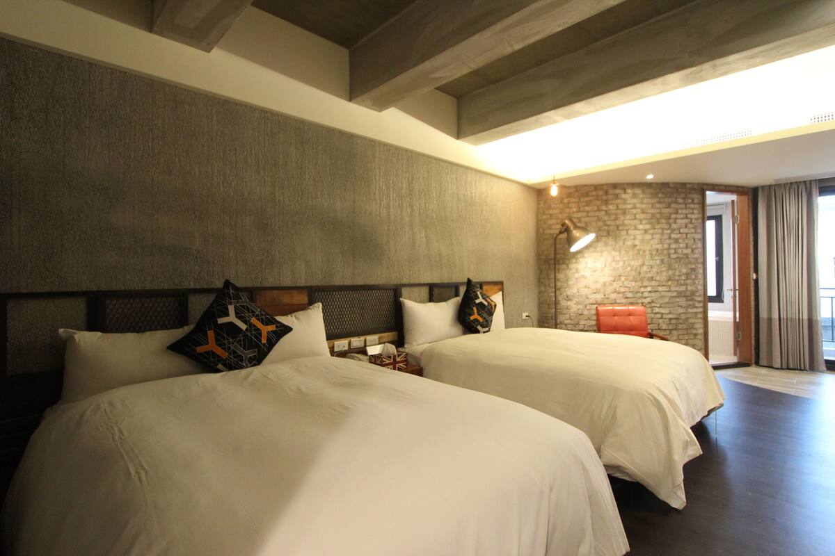 A hotel room with two beds, a stone wall, a modern lamp, and a view of the outdoors.