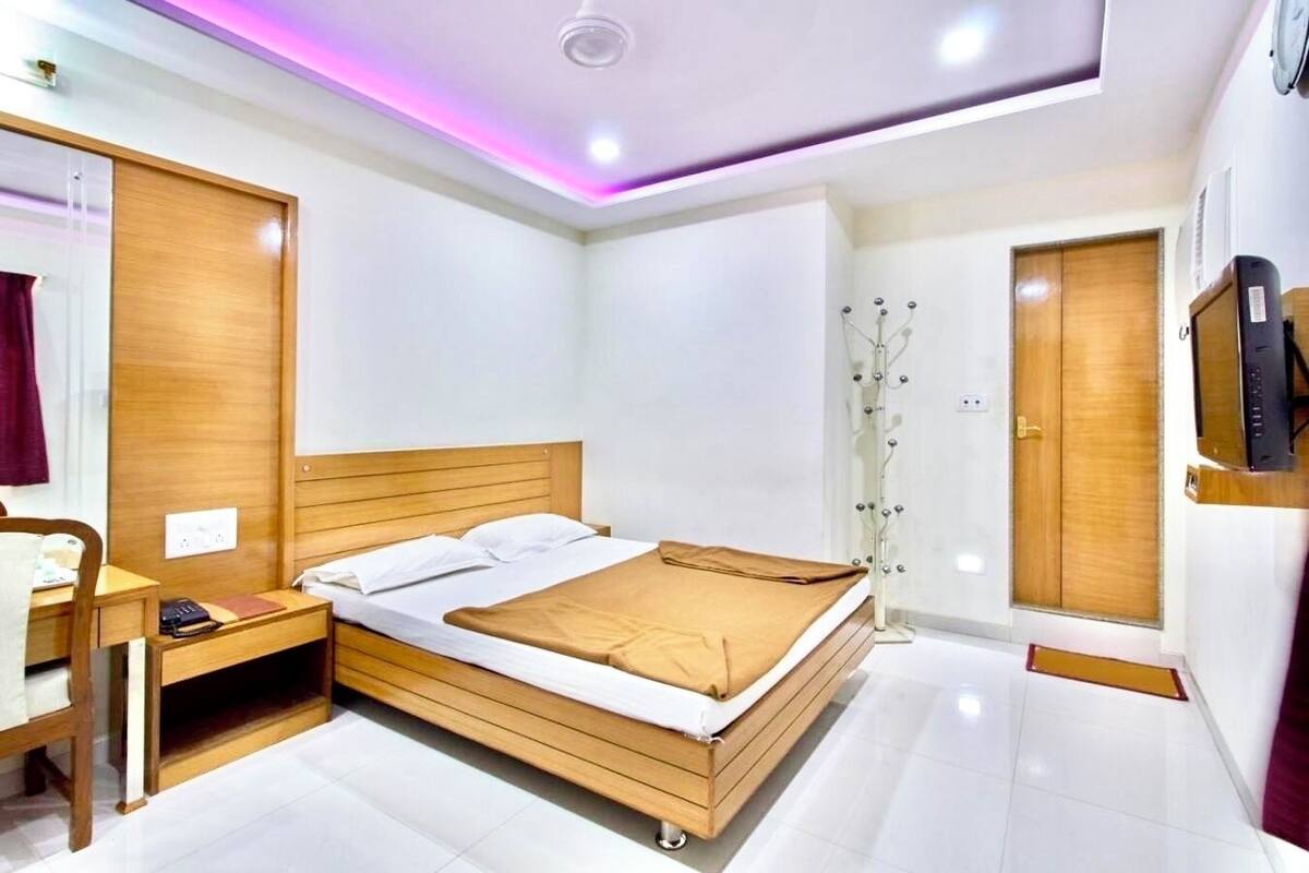 A modern bedroom with a wooden bed, a desk with a chair, a wardrobe, and a television mounted on the wall.