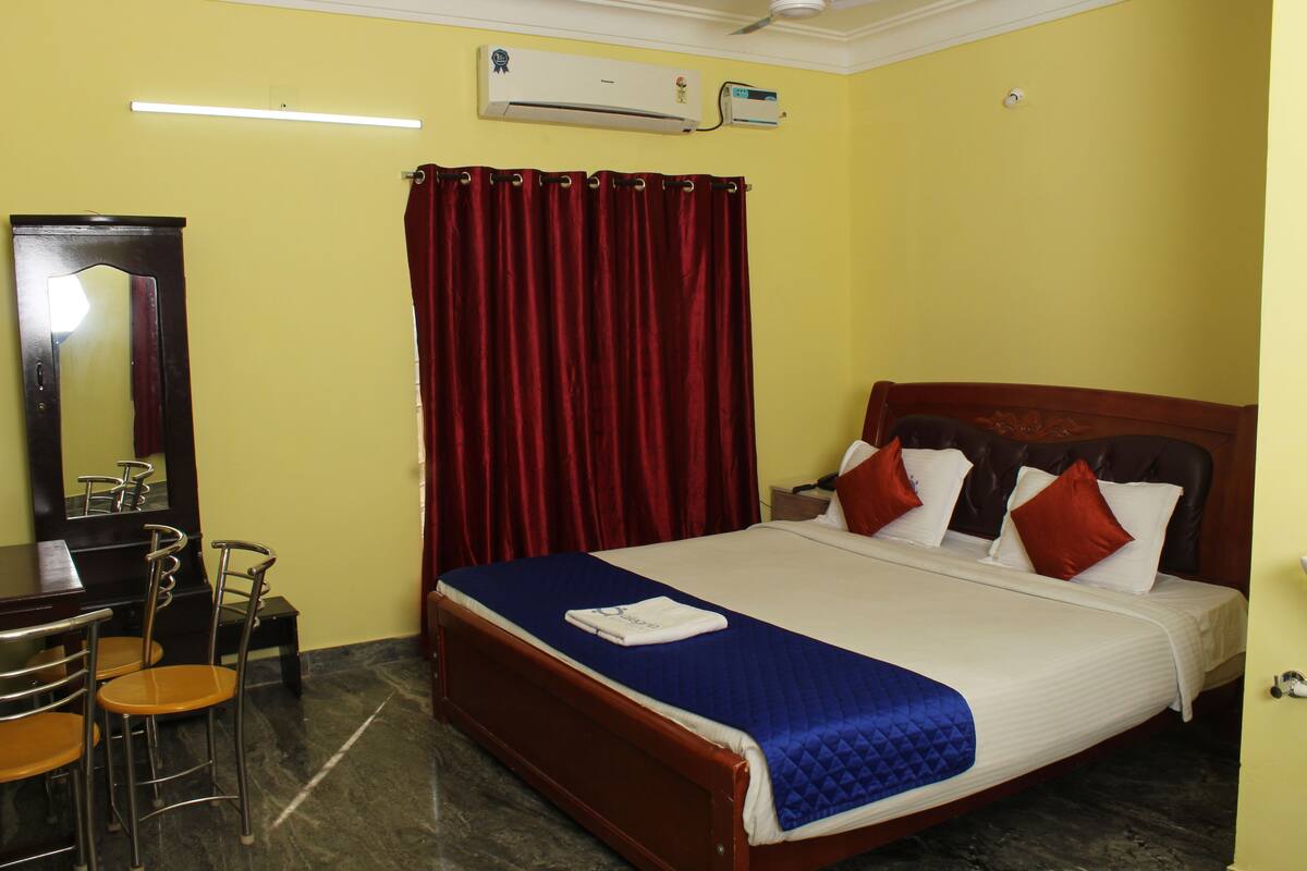 A hotel room with a bed, a desk, chairs, a mirror, and an air conditioning unit.