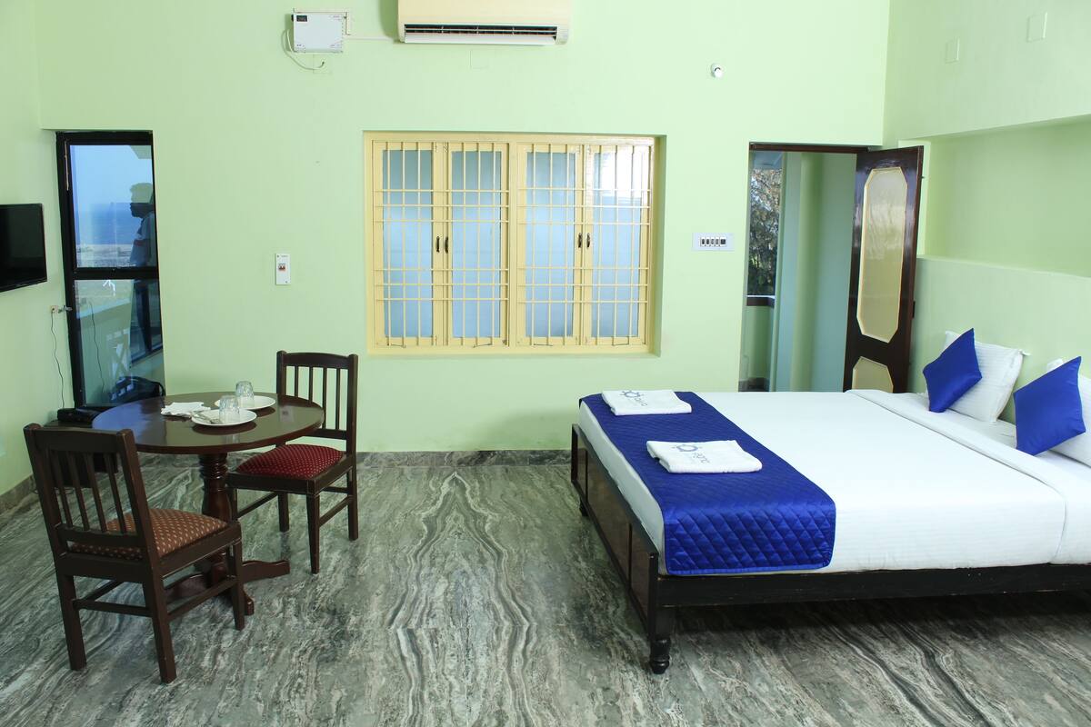 A hotel room with a bed, a dining table with chairs, a window with bars, and an air conditioning unit.