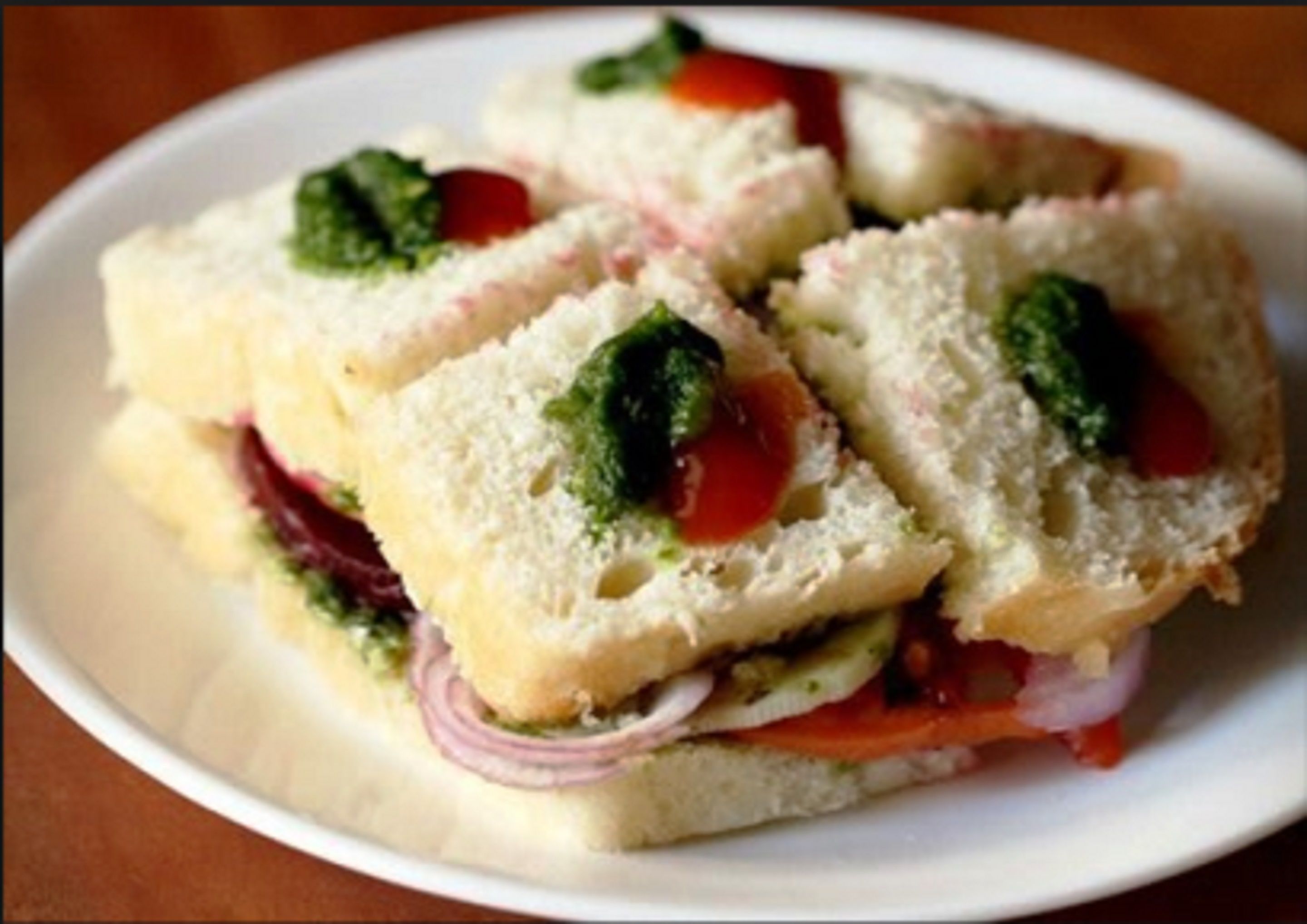 A plate with four sandwiches cut into squares, each with a slice of tomato, a leaf of basil, and a slice of red onion.