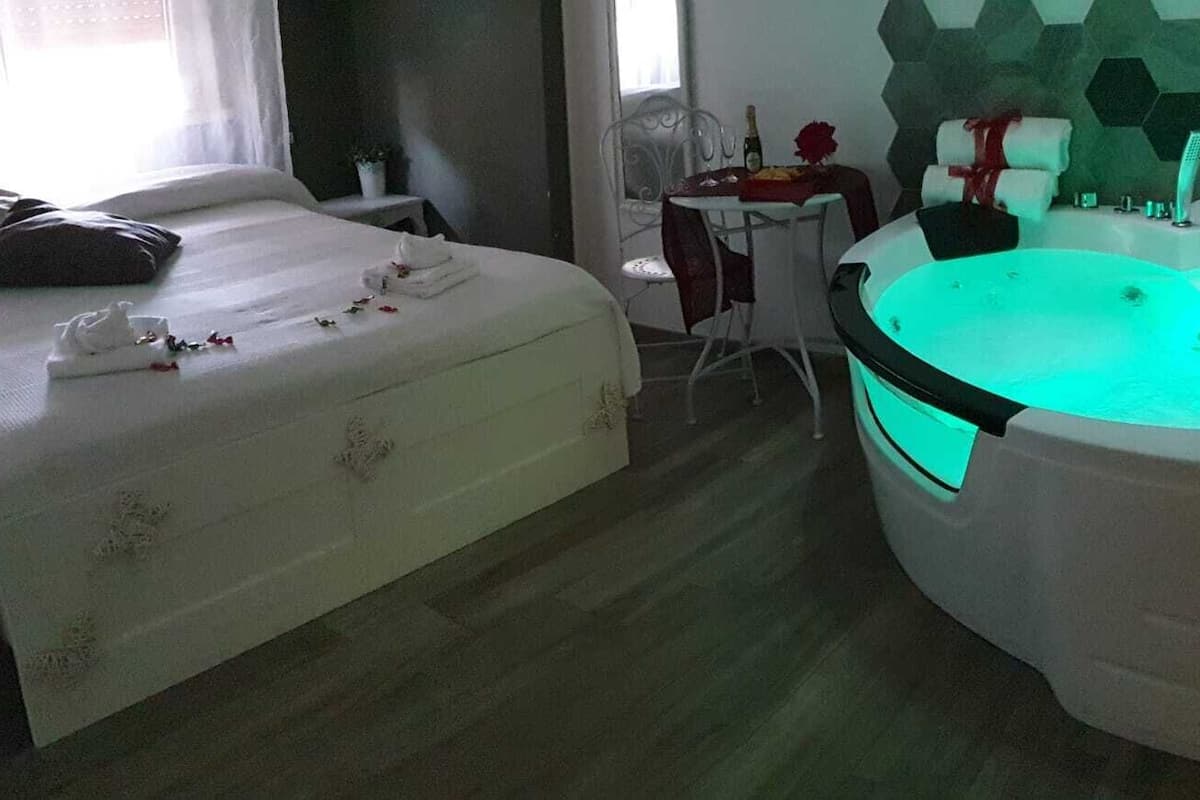 Habitación de hotel con cama, una mesita con una botella y flores, y una bañera grande con luz verde.
