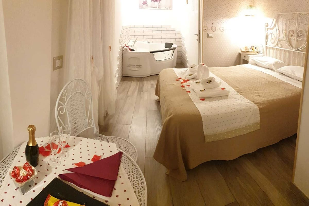 Una habitación de hotel con una cama, una mesita con una botella y vasos, y una silla.