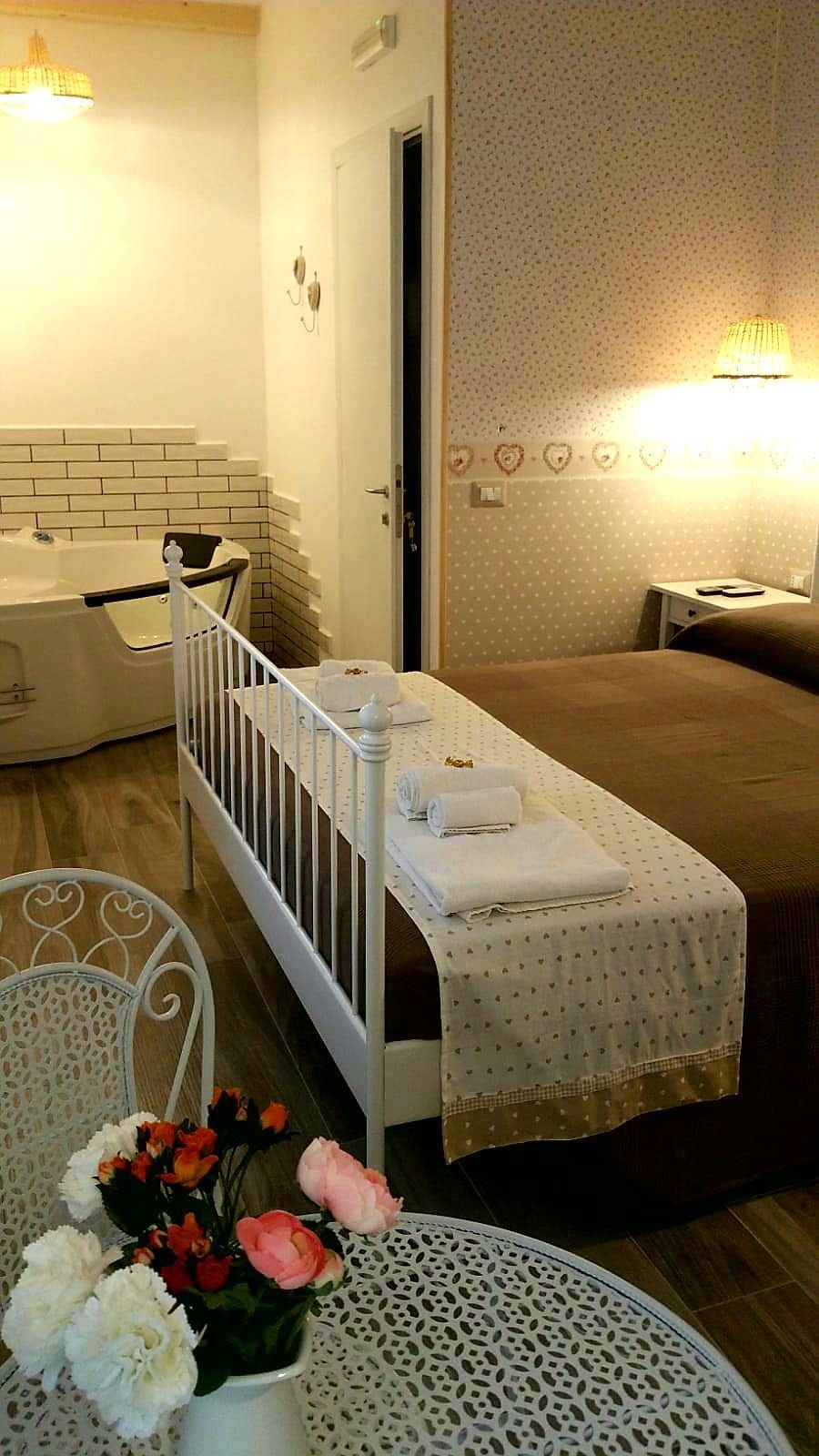 A hotel room with a white crib, a bed with a brown bedspread, a white table with a lace tablecloth, and a vase with flowers.