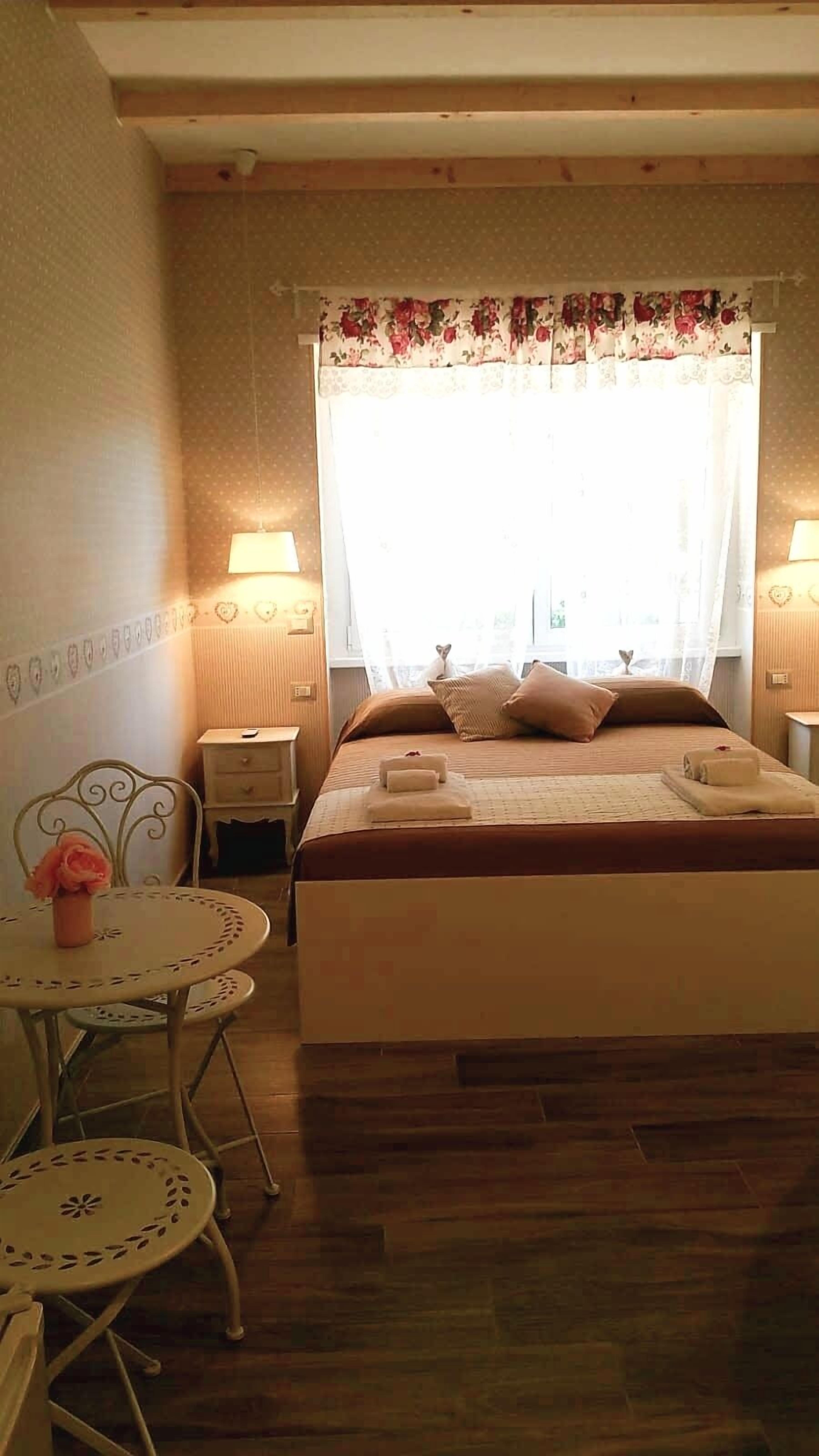 Un dormitorio con cama, mesitas de noche, una pequeña mesa redonda con sillas y una ventana con cortinas.