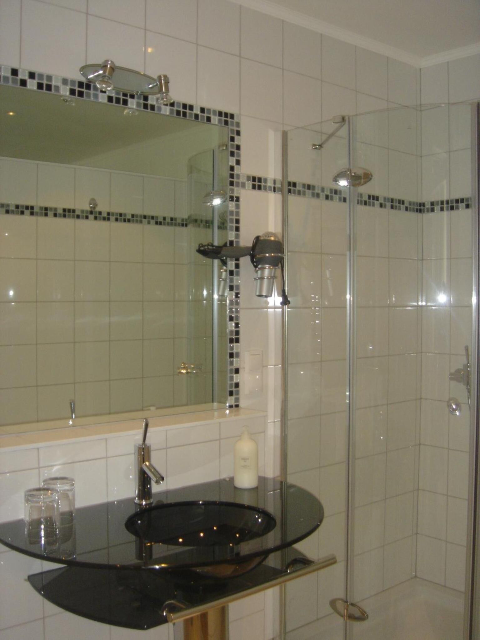 Ein Badezimmer mit einer Duschkabine, einem runden Waschbecken mit schwarzer Arbeitsplatte und einem Spiegel mit Mosaikfliesen.