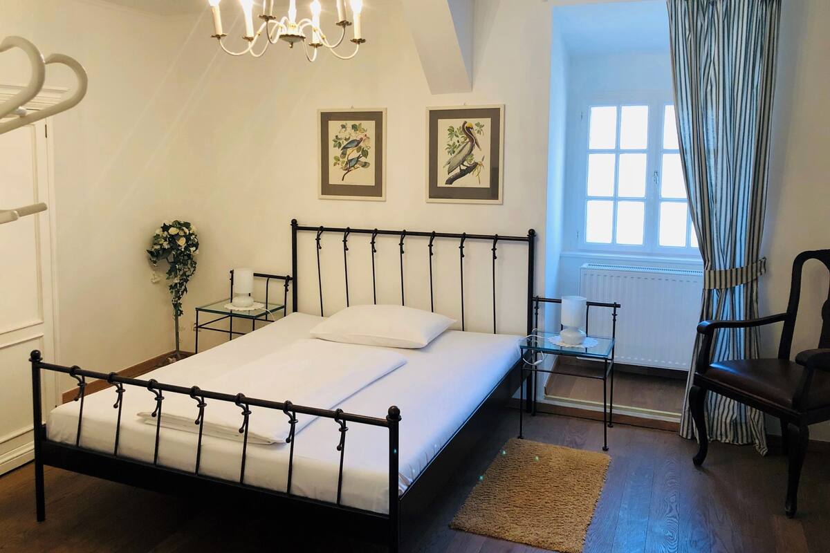 A bedroom with a black metal bed frame, white bedding, a chandelier, a window with curtains, and two framed pictures on the wall.
