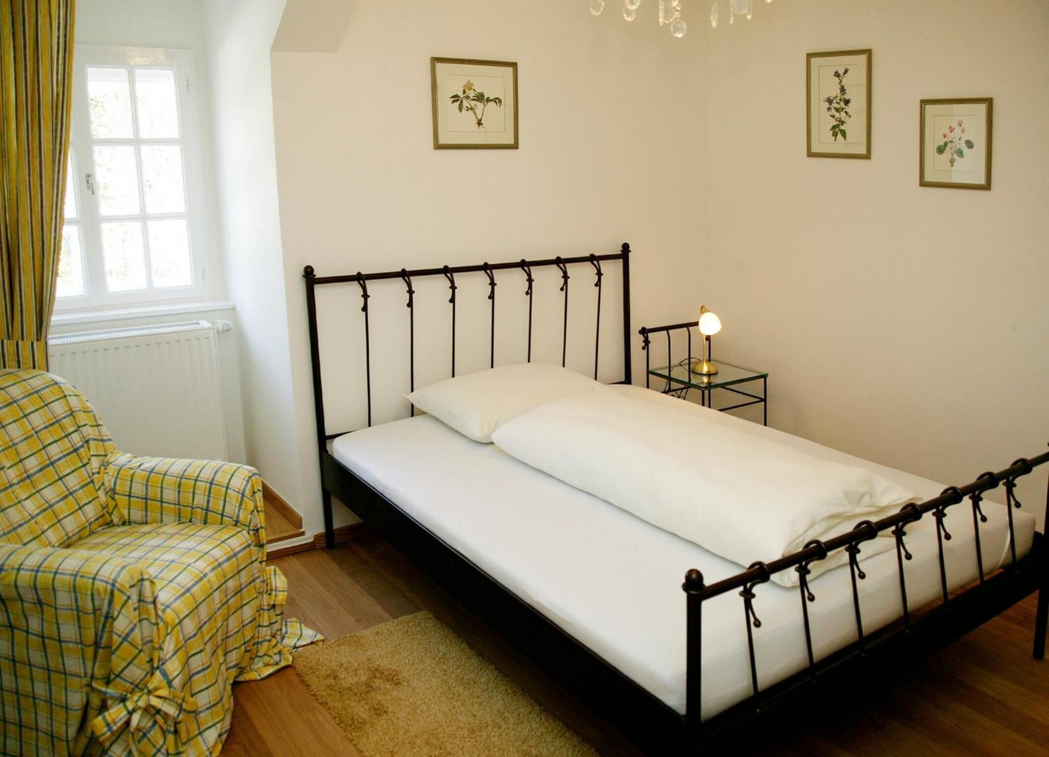 Uma cama de solteiro com estrutura de metal, uma mesa de cabeceira com abajur, uma cadeira com capa xadrez e impressões botânicas emolduradas na parede.