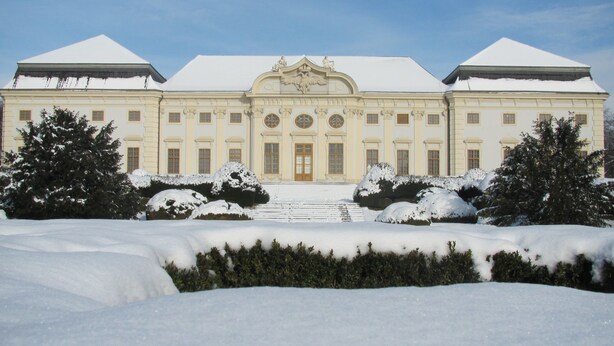 Un edificio grande y simétrico con una entrada central, flanqueado por árboles cubiertos de nieve y un césped nevado.
