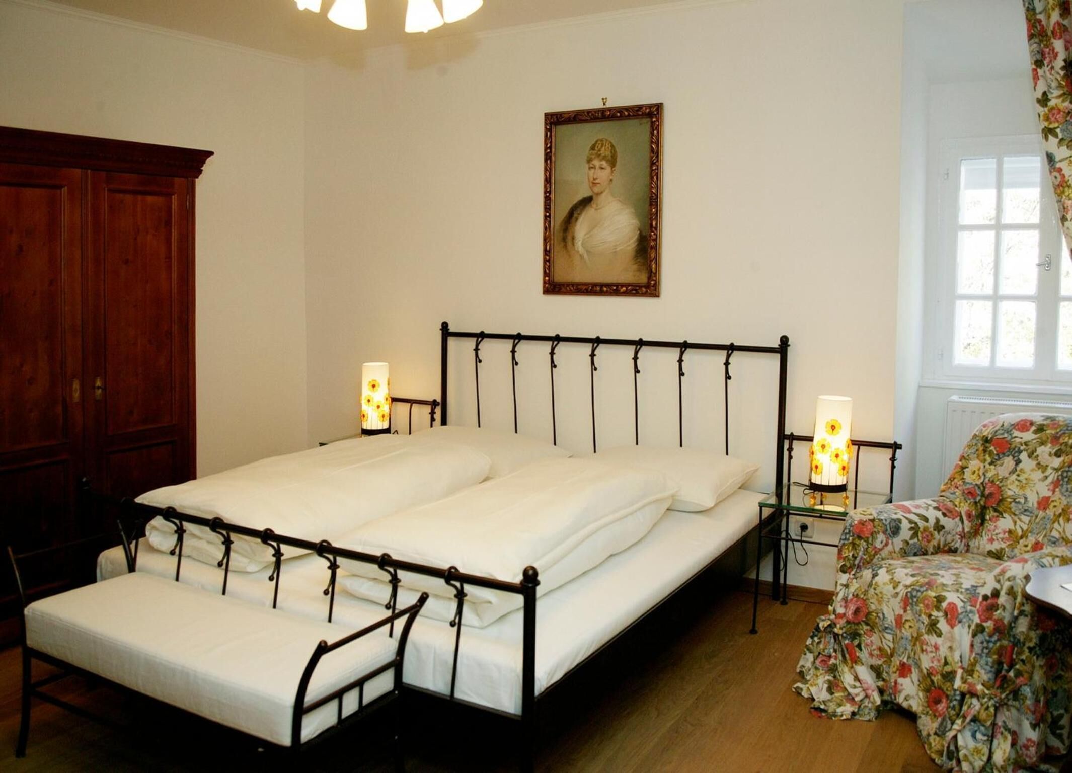 Un dormitorio con una cama doble, dos mesitas de noche con lámparas, un armario de madera, un sillón con estampado floral y un retrato en la pared.