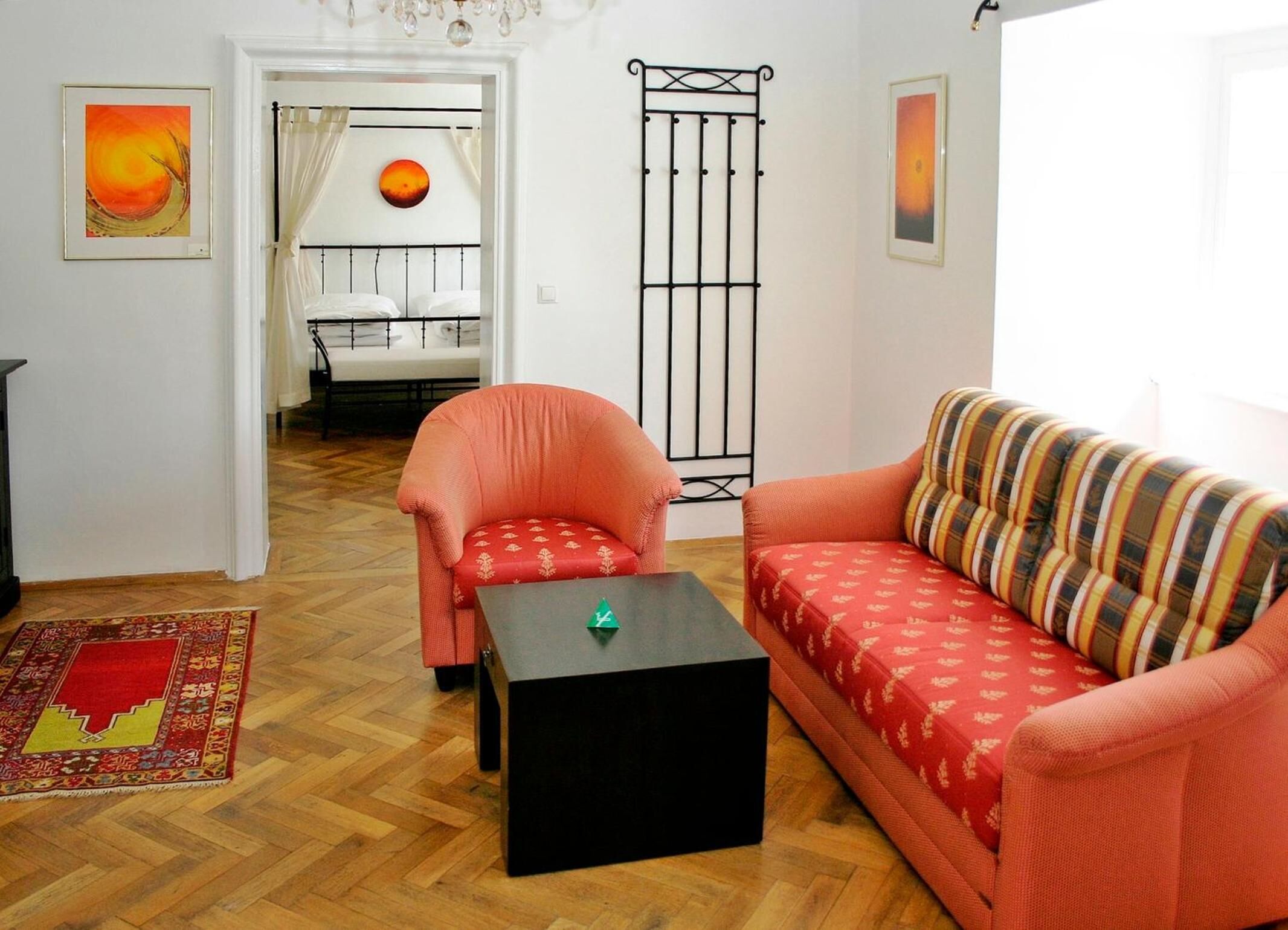 En stue med sofa, lænestol og et lille bord. Der er en seng i baggrunden med gardin og et panelradiator.