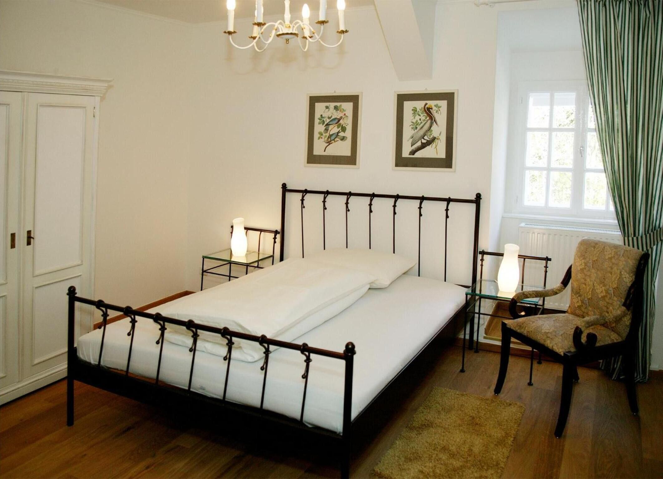 A bedroom with a four-poster bed, two bedside tables with lamps, a chair, and two framed bird paintings on the wall.
