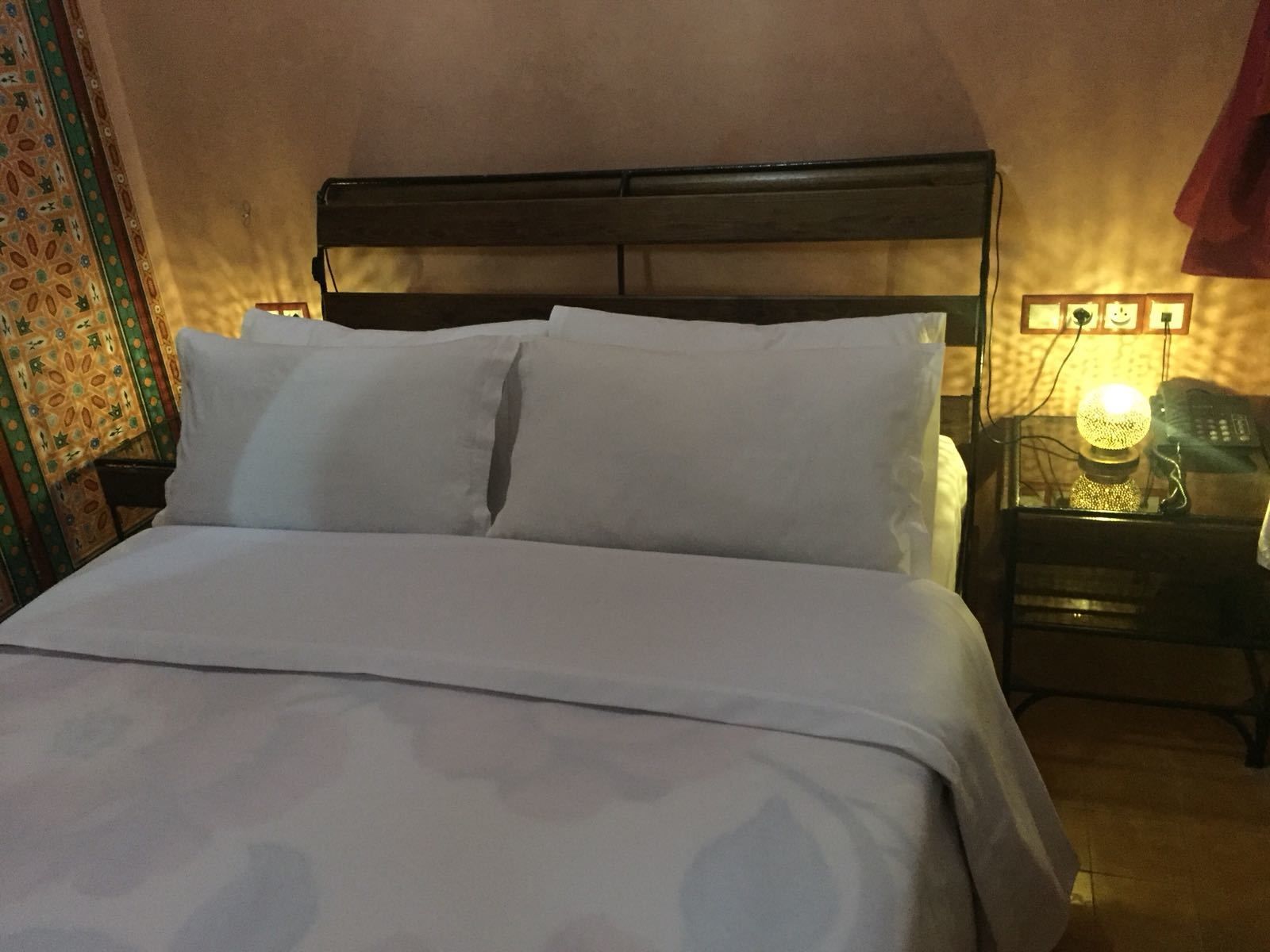 Una cama bien tendida con sábanas blancas y un cabecero de madera. Hay una mesita de noche con una lámpara y un teléfono.
