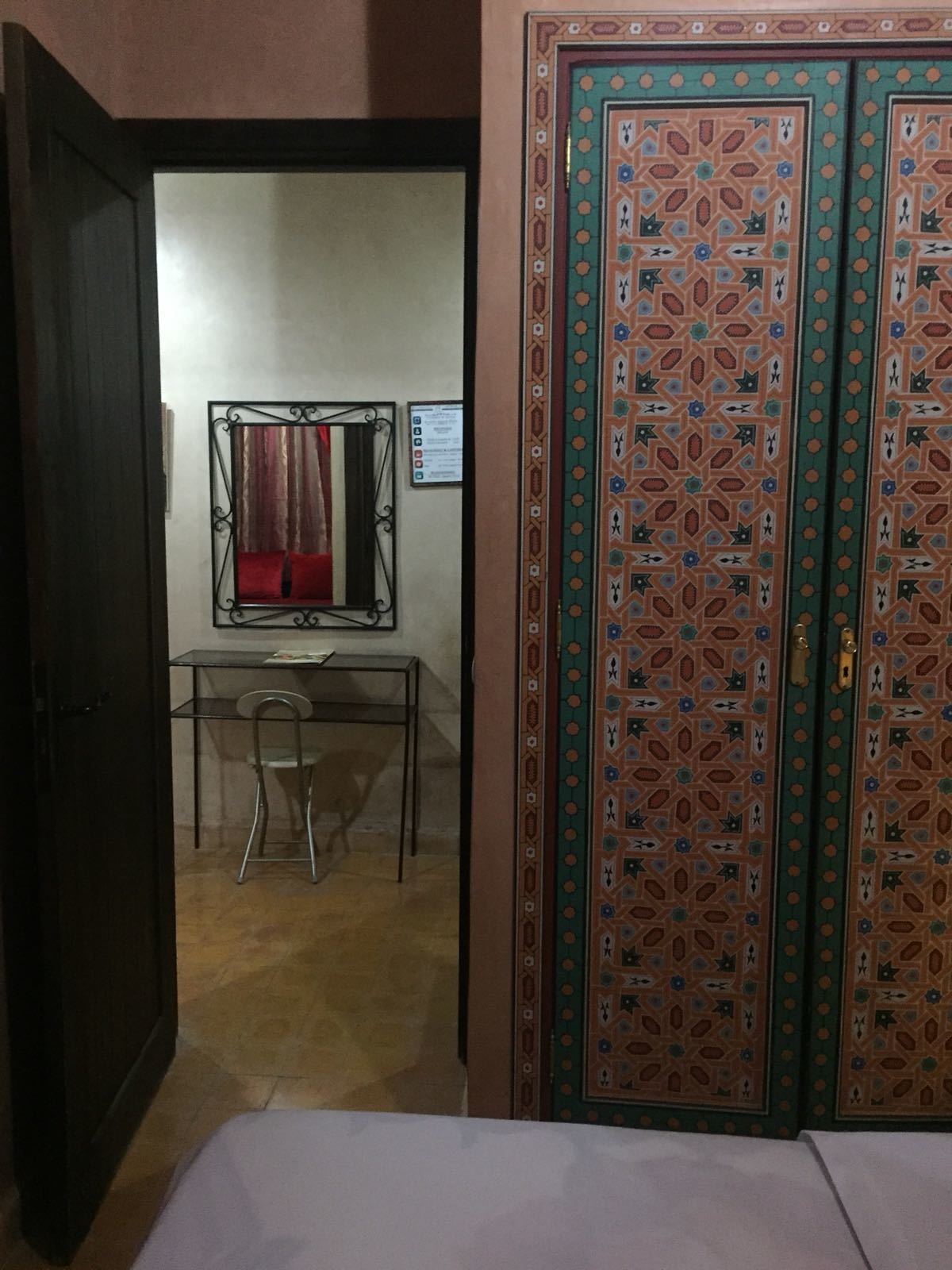 Ein Zimmer mit einem Bett, einem Schreibtisch mit Stuhl, einem Spiegel und einer Tür mit Muster.